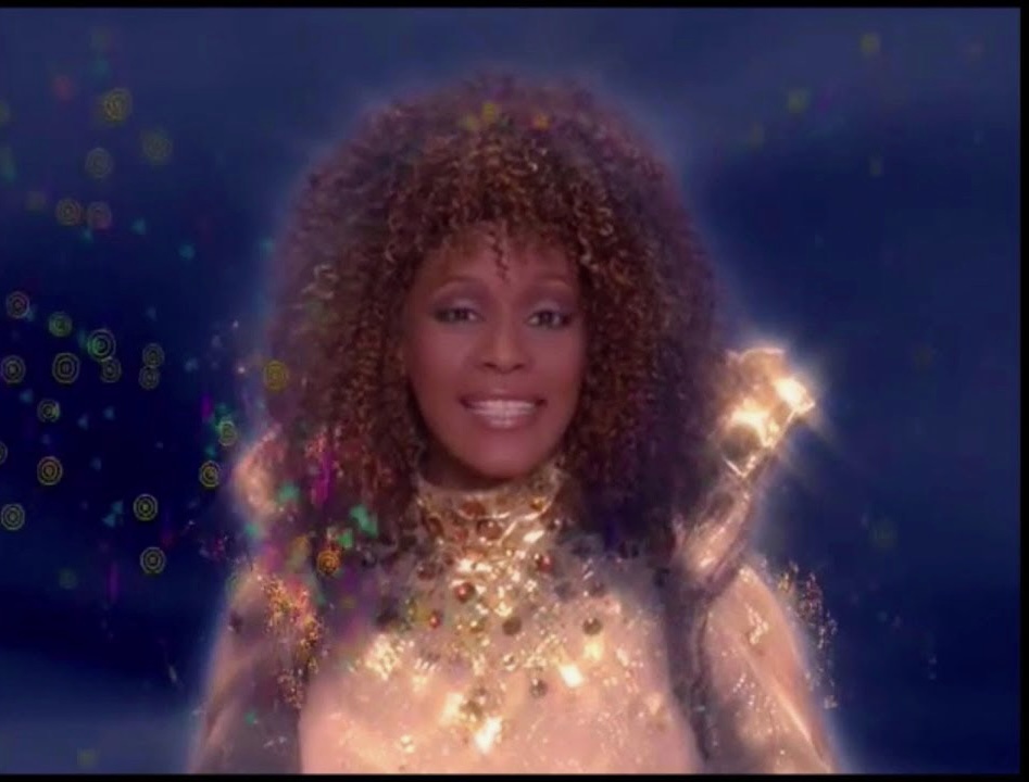 Whitney Houston in character as the Fairy Godmother in Rodgers and Hammerstein's Cinderella. She wears a gold gown with shining metallic accents. There are tiny dust-like particles floating around her face. She smiles. The indigo background behind her recalls an enchanted the kind of enchanted evening backdrop described in fairy tales.
